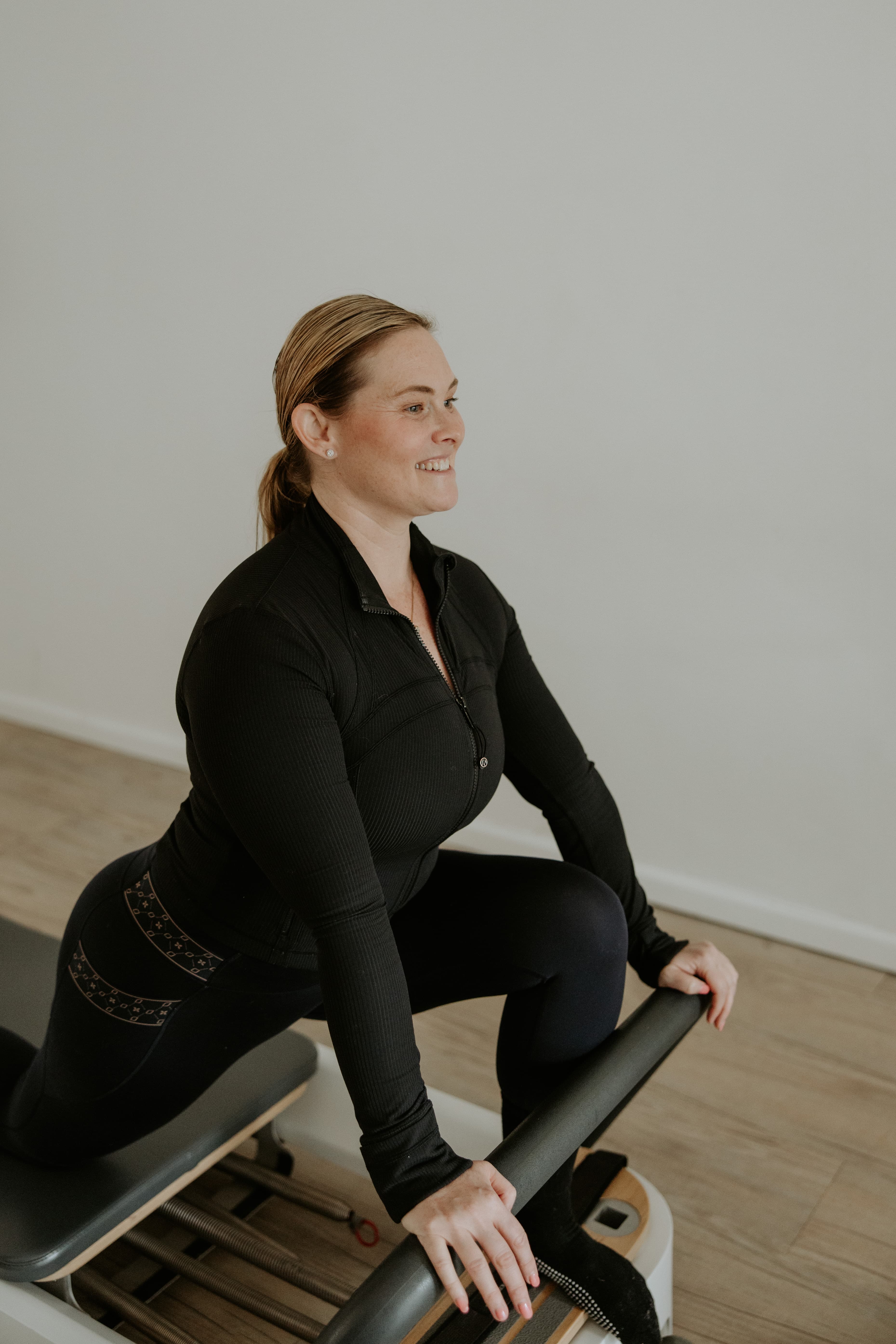 Smiling woman in black athletic wear performs a lunge stretch on a Pilates reformer.