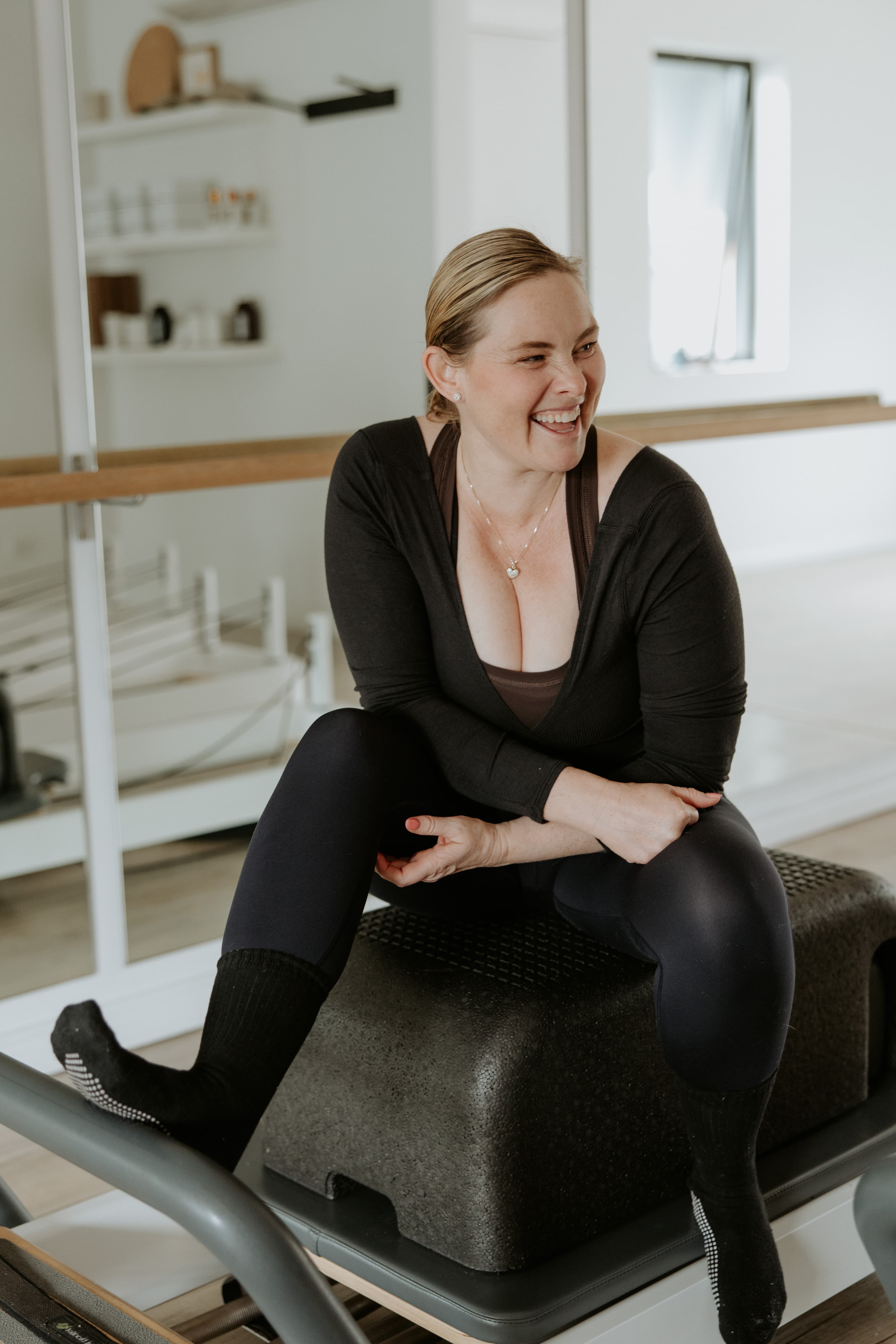 Laughing woman in black activewear sits on a Pilates reformer box in a bright studio.