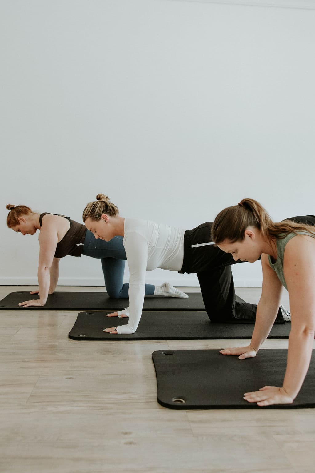Three women on yoga mats performing bird-dog exercises in a bright, minimalist fitness studio.