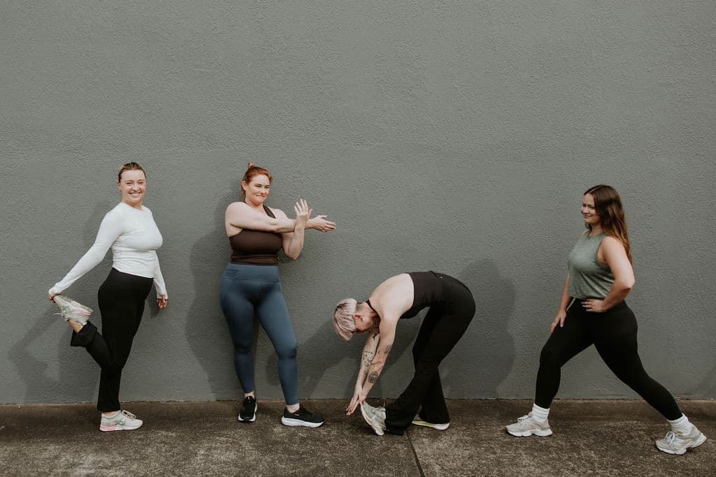 Four women in athletic wear performing different stretches against a textured grey wall.