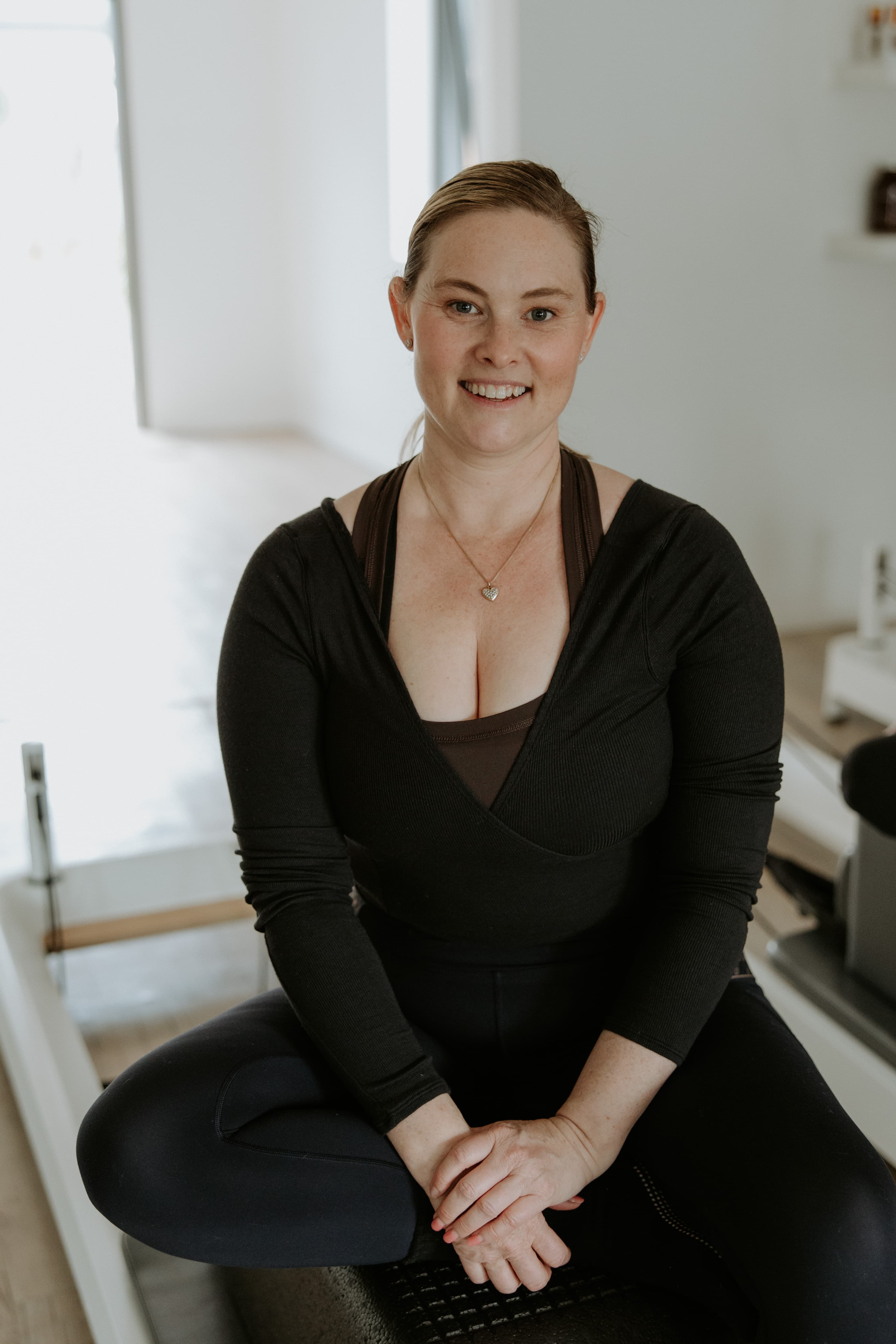 Smiling woman in black activewear sits cross-legged on a Pilates reformer in a studio.