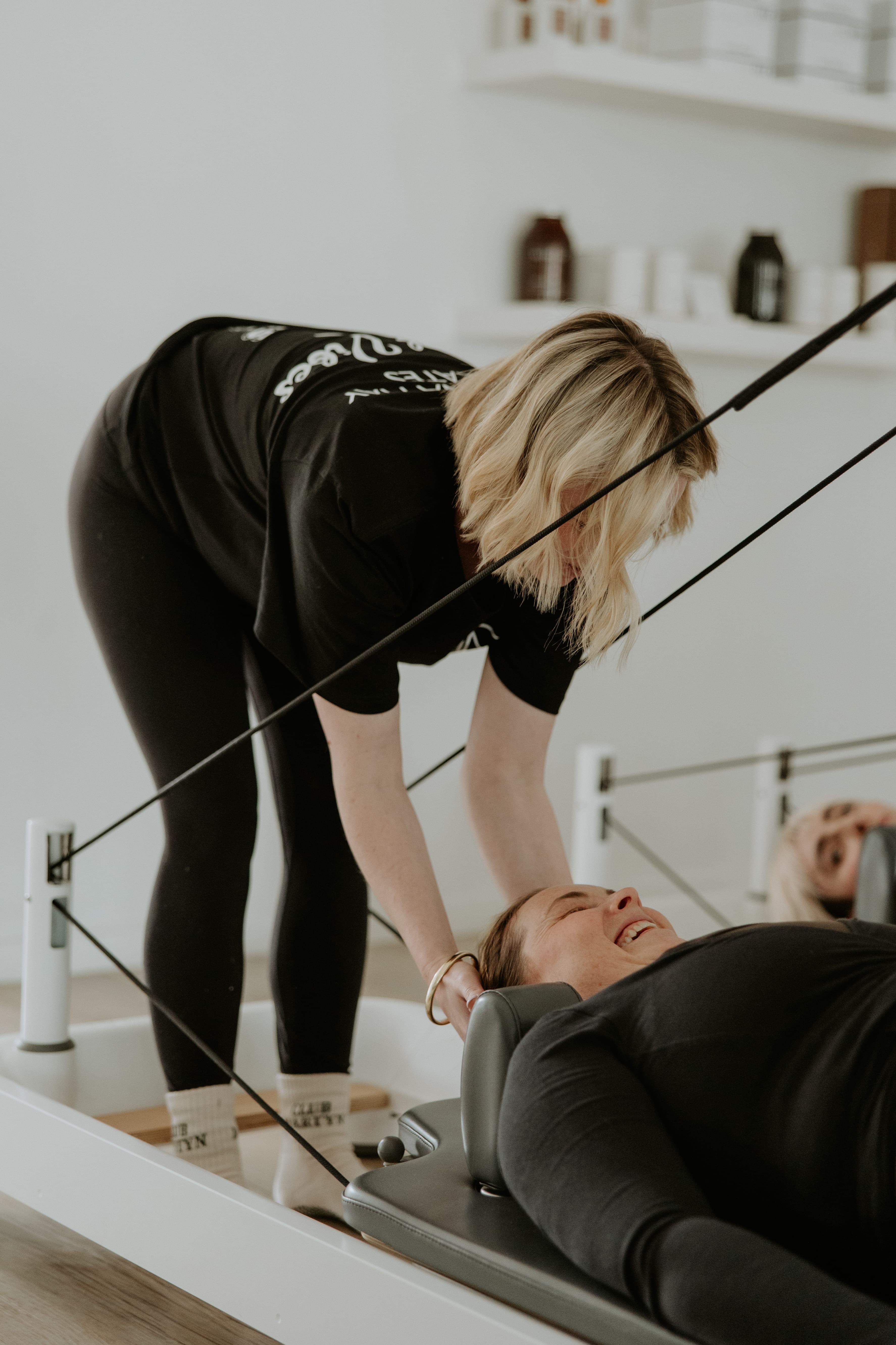 Blonde instructor adjusts a smiling woman's head position on a white Pilates reformer machine.