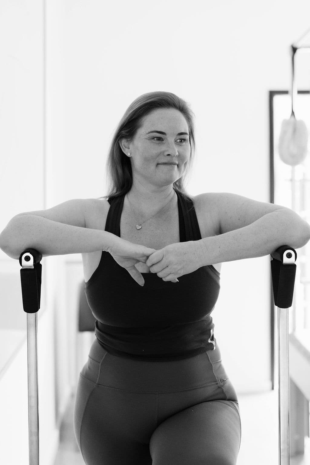 Smiling woman in athletic wear leaning on gym equipment in a black and white studio portrait.