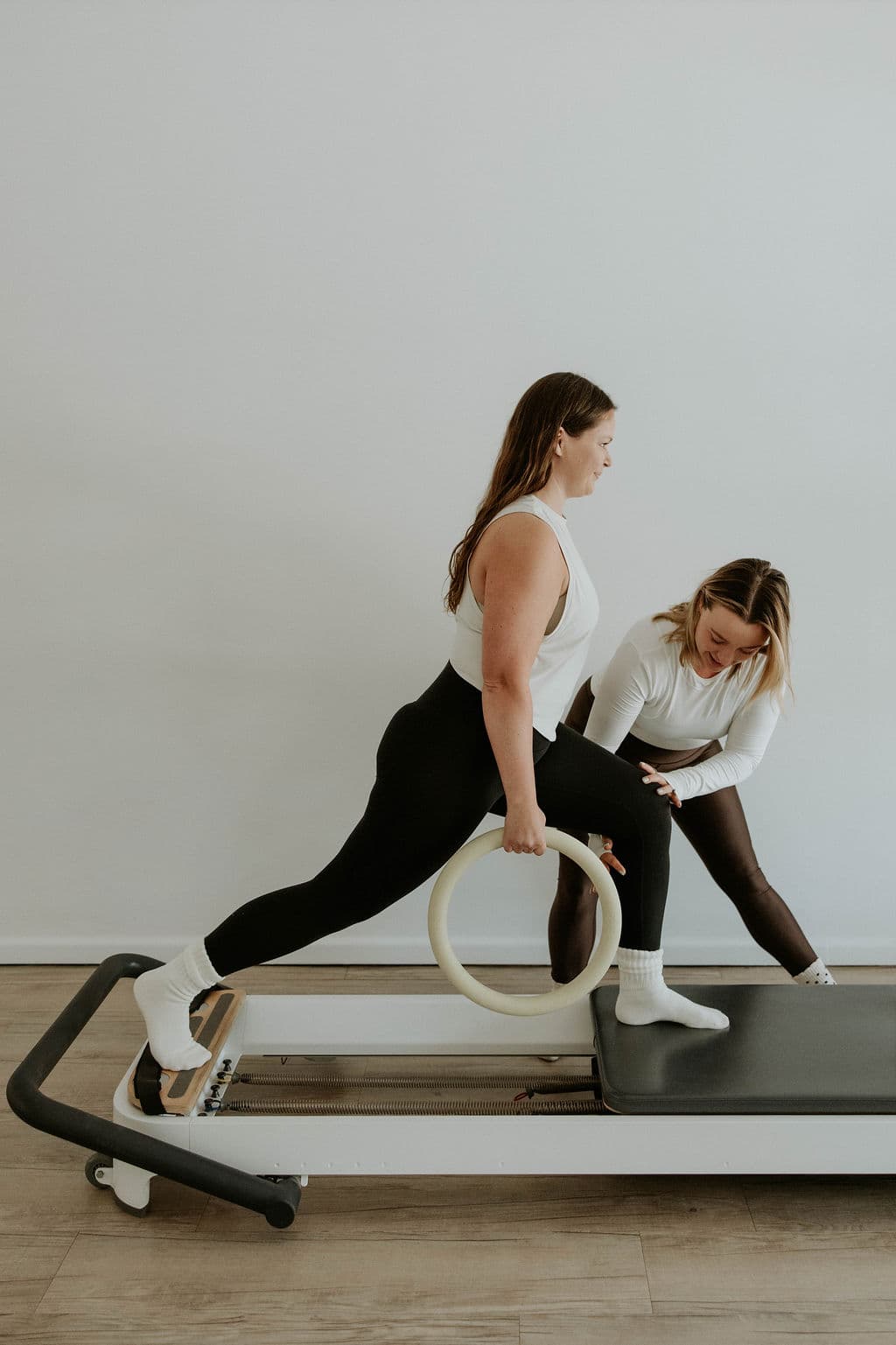 Woman holding a Pilates ring while performing a lunge on a reformer with an instructor.