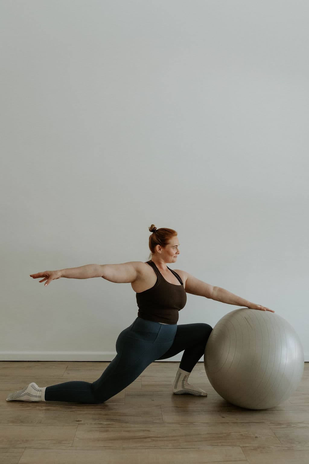 Red-haired woman in activewear performs a lunge stretch using a large grey exercise ball.